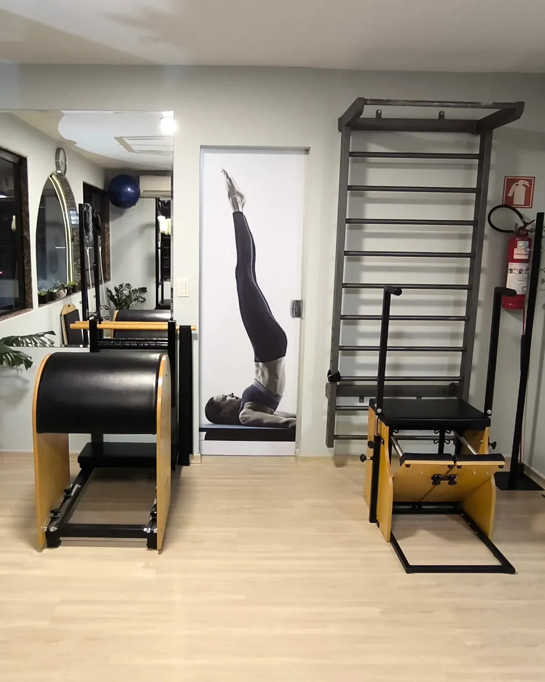 A sala do estúdio, vazia: um barrel de madeira e couro preto, a porta com um painel de pilates, o espaldar na parede e uma chair, sobre piso claro.